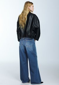 Černá kožená krátká bunda s nadměrným designem v kombinaci s volnými modrými džíny s širokými nohavicemi a vybledlým efektem. Má velké zadní kapsy.