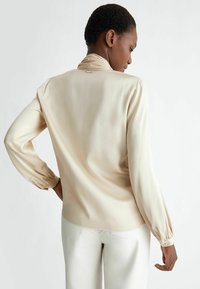 Blusa a maniche lunghe di un pallido beige, con una texture liscia, colletto alto, polsini arricciati e un piccolo accent metallico sul retro. Vestibilità facile.