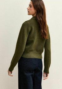 Mujer con cabello largo y marrón, vestida con un suéter de punto verde oliva y pantalones azul oscuro, de pie de lado contra un fondo liso.