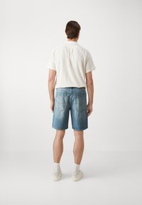 Gai krāsas īsām piedurknēm krekls, zili denima šorti un baltas sporta kedas ar teksturētu audumu un klasisku piegriezumu, skatīts no aizmugures.