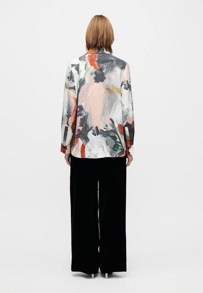 Monivärinen abstrakti kuvioitu paita kevyestä kankaasta, jossa on pitkät hihat ja napilliset kalvosimet; yhdistetty leveälahkeisiin mustiin housuihin.