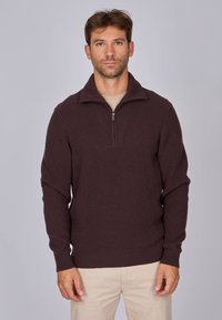 Pull en tricot marron foncé avec un col zippé, texture côtelée et manches longues, porté par-dessus une chemise beige plus claire.