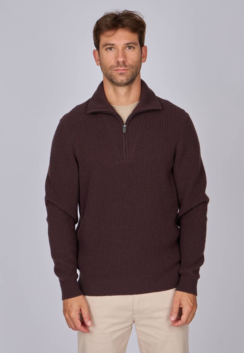Pull en tricot marron foncé avec un col zippé, texture côtelée et manches longues, porté par-dessus une chemise beige plus claire.