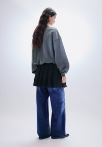 Ylimitoitettu harmaa collegepaita, jossa on lasketut olkapäät, kerroksittain mustan laskostetun hameen päällä, yhdistettynä leveälahkeisiin farkkuihin ja mustiin kenkiin.