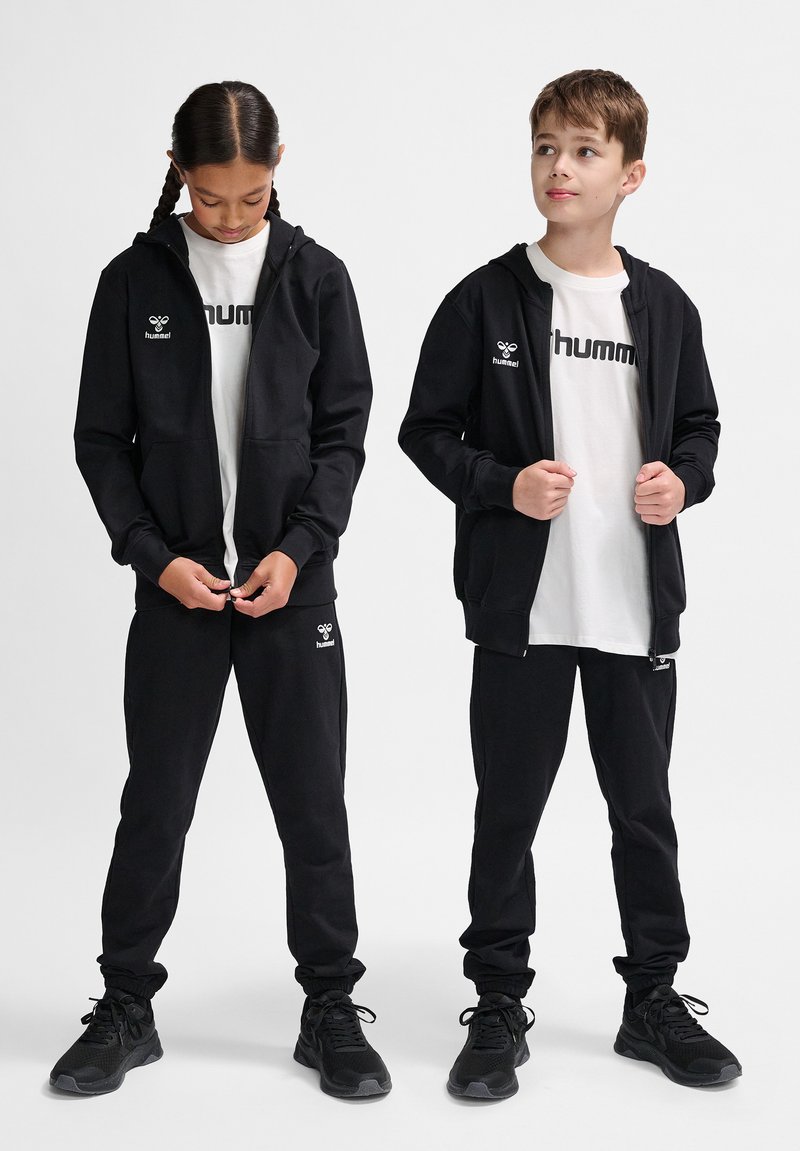 Schwarze Kapuzenjacken und -hosen, kombiniert mit weißen T-Shirts, auf denen "hummel" steht. Das Material wirkt weich, mit elastischen Bündchen und standardmäßiger Passform.