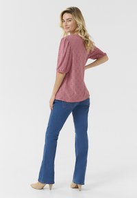 Blusa rosa con maniche a sbuffo, con motivi ad occhiello. Indossata con jeans azzurri svasati e scarpe con tacco color cammello, con il corpo leggermente inclinato all'indietro.
