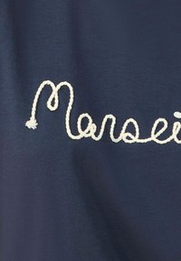 Tamsiai mėlynas audinys, kuriame yra išsiuvinėtas baltas kursyvinis tekstas „Marseille“. Paviršius lygus su minkštu apdailu.