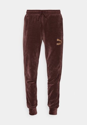 Personne portant un sweat à capuche et un pantalon en velours marron assortis Puma avec un logo doré Puma sur la poitrine et la cuisse, mains dans les poches.