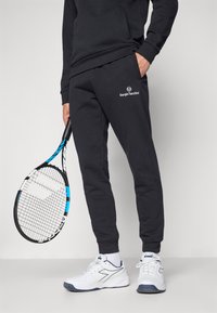 Fekete sportnadrág a Sergio Tacchini logójával, fehér teniszcipővel párosítva, kezében egy teniszütővel. Egyszerű dizájn és kiváló illeszkedés.