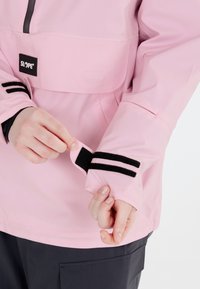 Rosa wasserdichte Jacke mit einer Fronttasche, grauem Reißverschluss und schwarzen Streifendetails an den Ärmeln sowie verstellbaren Klettverschluss-Manschetten.