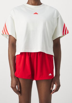 Person trägt ein weißes, kurzes T-Shirt mit roten Streifen an den Ärmeln und rote Sportshorts mit weißem Logo und steht vor einem schlichten Hintergrund.