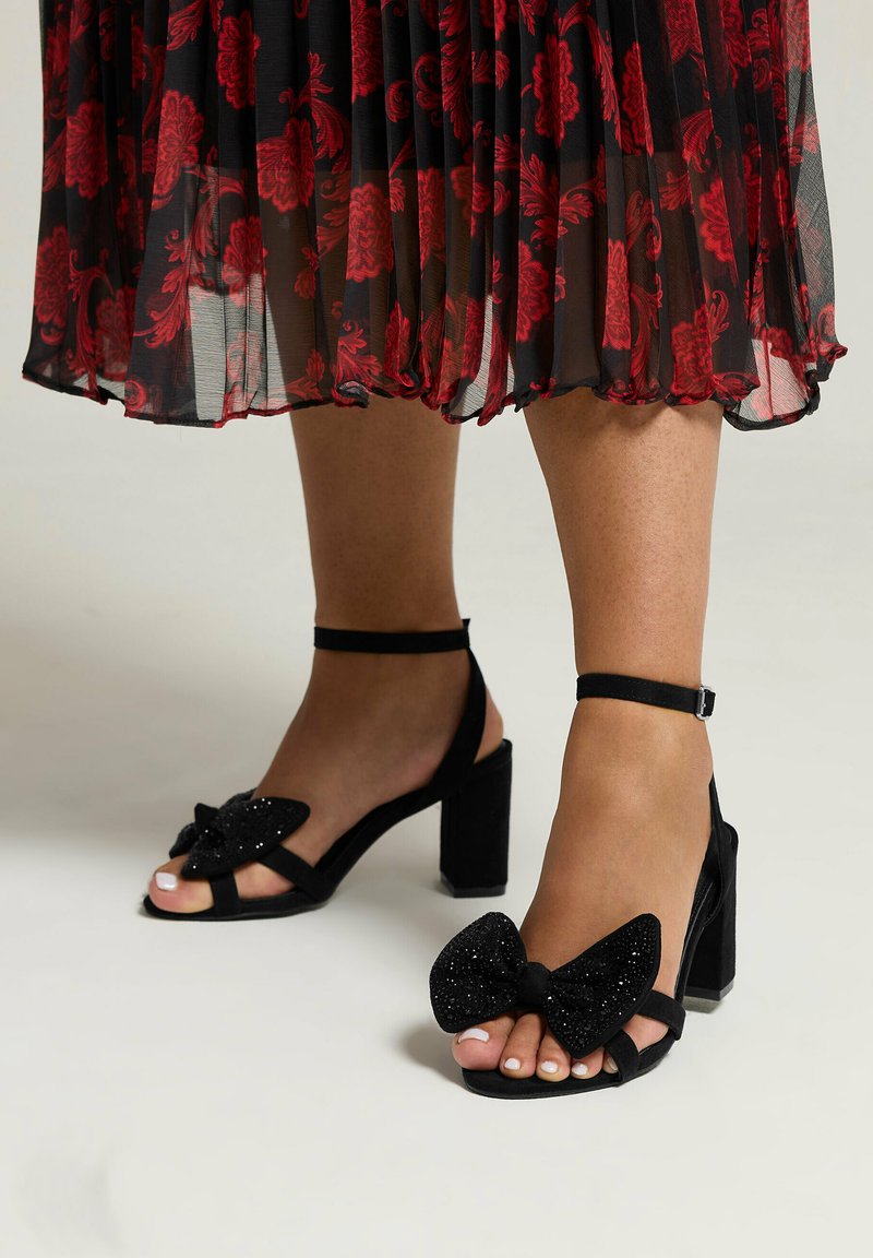 Black high-heeled sandals with a chunky heel, featuring a large black bow and shimmering accents. Strap around the ankle for support.