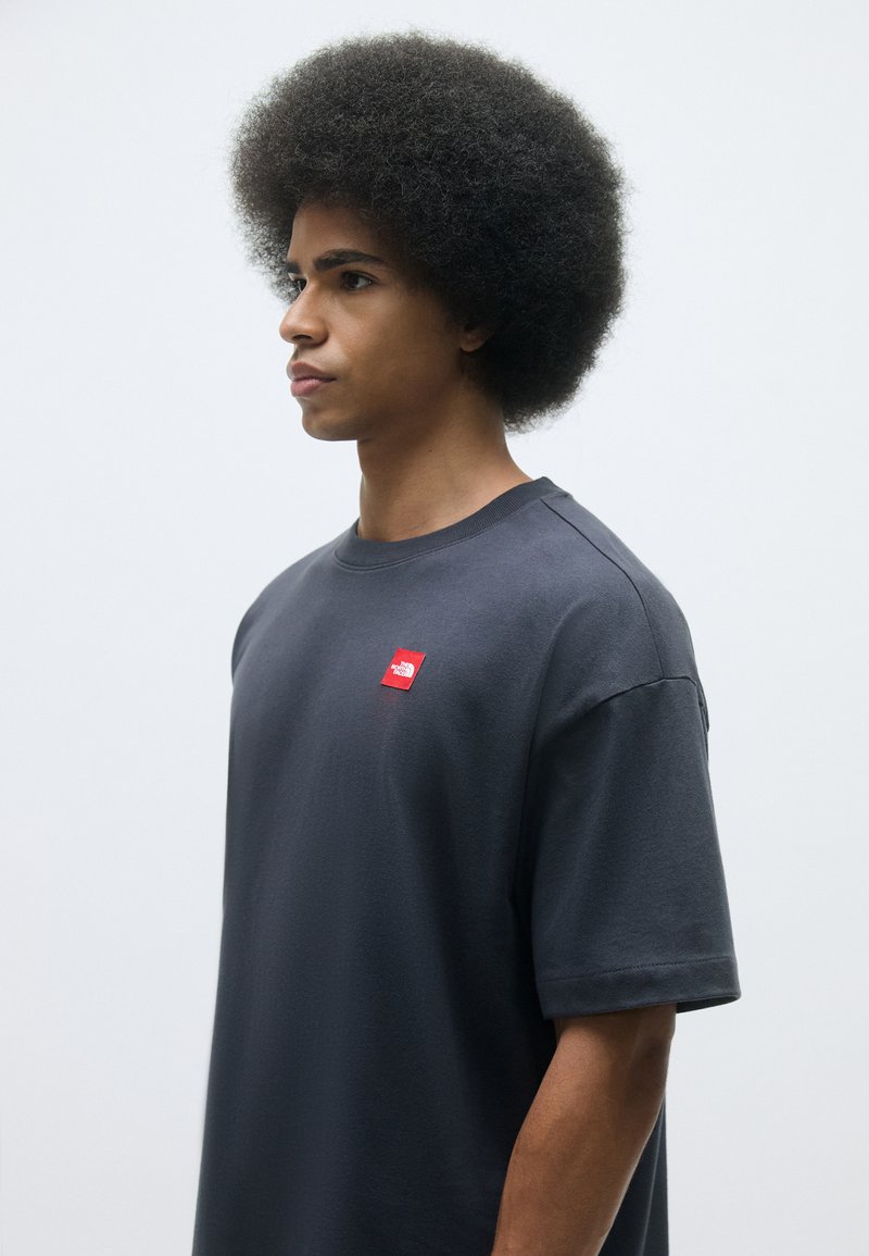 T-shirt oversize grigio scuro a maniche corte, con una piccola etichetta rossa sul petto. Tessuto morbido.