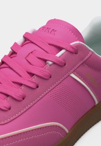 Κοντινό πλάνο ροζ αθλητικού παπουτσιού με υφασμάτινους κορδόνια, διάτρητη πλευρική επιφάνεια, λευκή επένδυση και καφέ σόλα, με το λογότυπο "ARKK" στη γλώσσα και την πλευρά.