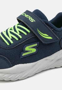 Tumes sinise võrgu ja säravroheliste aktsentidega spordijalu, millel on rihma sulgemine, tekstureeritud põhi ja logo detail.
