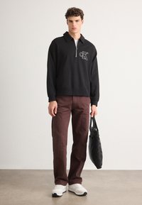 Maglione nero con zip e logo ricamato, pantaloni bordeaux e sneakers bianche. Il modello tiene una borsa tote nera, in piedi su un pavimento liscio.