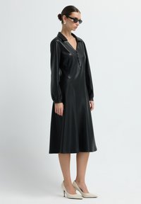 Žena v černých kožených midi šatech s dlouhým rukávem, bílé vysoké podpatky, černé sluneční brýle a stříbrné náušnice, stojí na bílém pozadí.