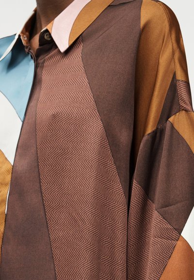 Lähikuva monivärisestä, kuvioidusta paidasta, jossa on ruskeita, sinapinkeltaisia, vaaleansinisiä, valkoisia ja pinkkejä kangaspaneeleja ja geometrinen siksak-kuvio.