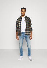 Jovem vestido com uma camisa xadrez preta e amarela sobre uma t-shirt branca com estampado, jeans de tom azul claro e ténis brancos, encostado a uma parede branca lisa.