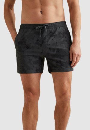 Mężczyzna noszący czarne kąpielówki w wzory z troczkiem w talii, stojący z rękami blisko kieszeni na jednolitym tle.