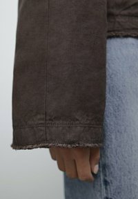 Handen avslappnad vid sidan, iklädd en brun långärmad skjorta med fransade manschetter och ljusblå denimjeans.