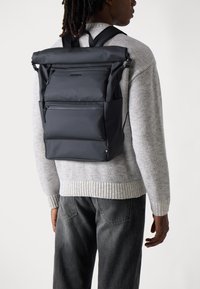 Black roll-top backpack made from smooth material, featuring multiple zipped pockets and adjustable straps, worn over a gray sweater.