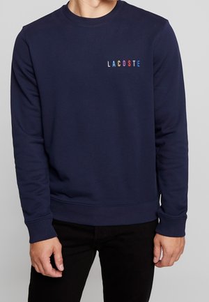 Mann trägt dunkelblauen Pullover mit mehrfarbigem "LACOSTE"-Schriftzug auf der Brust und schwarze Hose, steht vor einfachem Hintergrund.