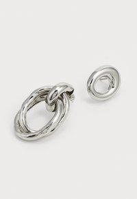 Pendientes de metal plateado con una forma ovalada entrelazada y un pequeño diseño circular, ambos con un acabado brillante y pulido.