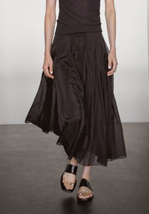 Person wearing a flowing dark skirt and sleeveless top paired with black open-toe sandals walking on a plain floor against a light wall.