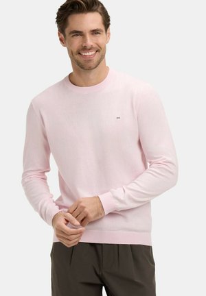 Homme souriant portant un pull à manches longues rose clair et un pantalon foncé, debout devant un fond blanc uni.