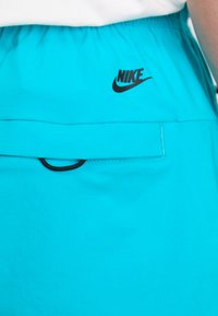 Turkise træningsshorts med et sort Nike-logo, en elastisk talje og en lille sort lomme-design detalje. Glat stoftekstur.