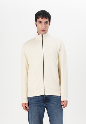 Light beige zip-up jacket with a high collar, made from soft material. Features a subtle logo and ribbed cuffs, paired with blue jeans.