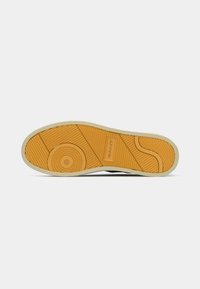 Gummisohle eines Sneakers in einer kontrastierenden gelb-braunen Farbe. Verfügt über geriffelte Muster und einen kreisförmigen Logoabdruck mit einem glatten weißen Rand.