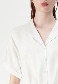 Ingrandimento di una persona che indossa una camicia bianca a maniche corte con colletto, mostrando le labbra, il collo e la parte superiore del petto.