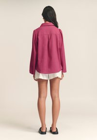Magenta langemouwen shirt met kraag, geplooide schouders en een achterste plooi, gecombineerd met witte shorts en zwarte instapschoenen.