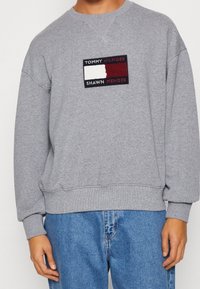 Šedá mikina vyrobená z bavlněné směsi s žebrovanými manžetami a lemem. Na přední straně má obdélníkový logotyp v červené, bílé a černé barvě. Pohodlný střih.