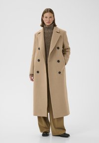 Cappotto doppiopetto in misto lana beige con grandi bottoni neri, rever a scollatura e tasche laterali. Abbinato a un dolcevita marrone e pantaloni beige.