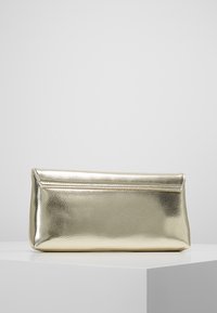 Obdélníková kovová zlatá kabelka s klopou, vystavená na bílé ploše před jednoduchým šedým pozadím.