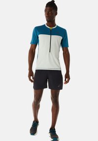 Atletische man in een blauw-wit shirt met halve rits, zwarte shorts en zwarte hardloopschoenen, staande tegen een effen witte achtergrond.