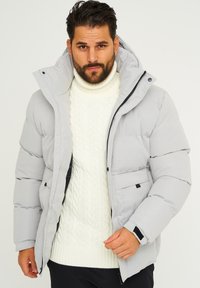 Hellgraue Daunenjacke mit hohem Kragen, Frontreißverschluss und Seitentaschen. Getragen über einem weißen Strickpullover für zusätzliche Wärme.