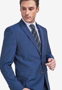 Blaues maßgeschneidertes Sakko mit Revers, Ein-Knopf-Verschluss und zwei Fronttaschen. Getragen über einem weißen Hemd und einer gestreiften Krawatte.