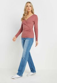Langärmliges Wickeloberteil in staubigem Rosé, mit V-Ausschnitt und gerafftem Seiten-Detail, kombiniert mit hellblauen, ausgestellten Jeans und weißen Sneakers.