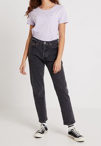 Camiseta de manga corta lila con un pequeño logo, combinada con jeans negros de cintura alta y zapatillas blancas y negras.