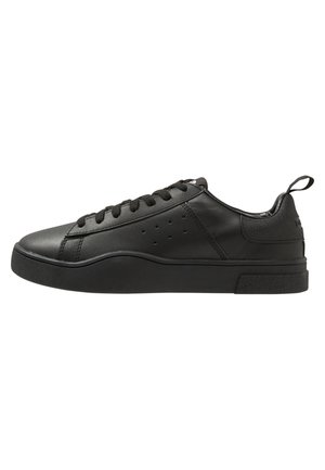 Zapatilla de cuero negro con puntera redondeada, detalles texturizados, perforaciones en el lateral, suela plana y una lengüeta en el talón.
