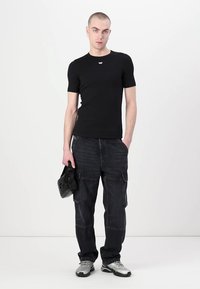 Svart kortermet t-skjorte med en liten logo nær nakken, kombinert med mørke cargo-bukser og grå joggesko. Modellen holder en svart veske.