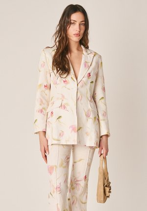 Mujer vestida con un traje floral crema que sostiene un bolso beige con un detalle de flor, de pie contra un fondo claro y liso.
