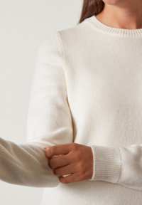 Maglione bianco lavorato a maglia con scollatura rotonda, tessuto testurizzato e polsini a coste. Il braccio sinistro è leggermente esteso, evidenziando la vestibilità.