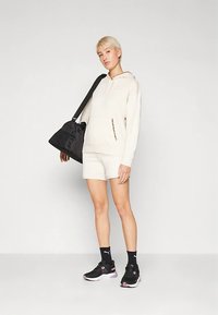 Sweatshirt bege com bolso frontal, combinado com calções bege. Ténis desportivos pretos com detalhes em rosa. Mala tote preta pendurada ao ombro.