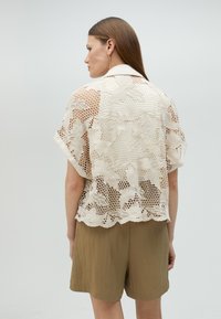Lace blouse in cream with floral pattern and open weave. Short sleeves, curved hem, and a collared neckline. Paired with olive shorts.