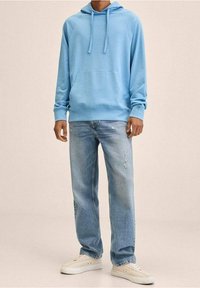Uomo con felpa blu chiaro, jeans blu sbiaditi con leggeri segni di usura e sneaker beige, in piedi contro uno sfondo semplice.
